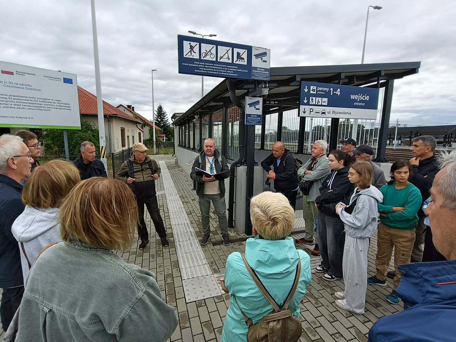 20260905-spacer-historyczny-dworzec-oswiecim-05 - NaszOświęcim - informacje i wydarzenia. Przewodnik czyta z notatek grupie uczestników przy wejściu na perony dworca kolejowego w Oświęcimiu