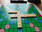 V Mistrzostwa Oświęcimia w Scrabble – grafika ilustracyjna - NaszOświęcim - informacje i wydarzenia. Plansza do Scrabble z ułożonymi słowami OŚWIĘCIM i ZAWODY, obok filiżanka kawy i stojak z płytkami, w tle sala turniejowa