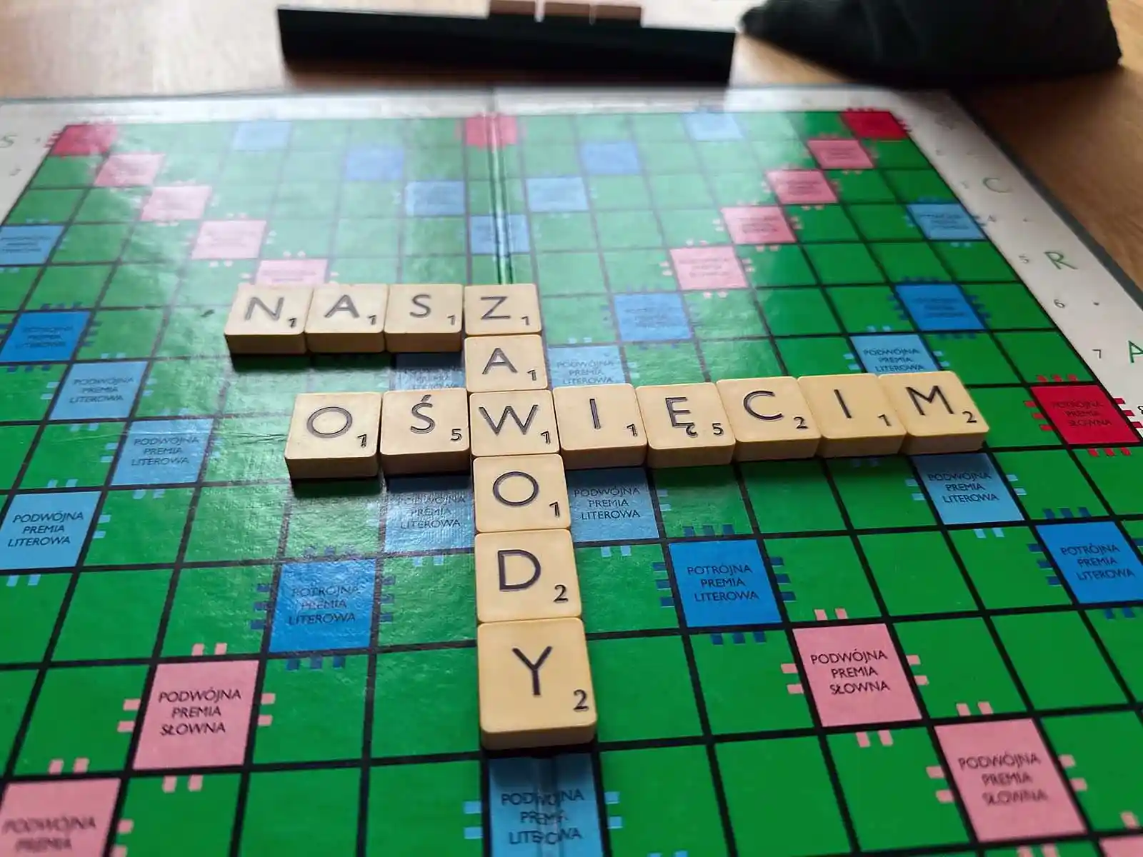 V Mistrzostwa Oświęcimia w Scrabble – grafika ilustracyjna - NaszOświęcim - informacje i wydarzenia. Plansza do Scrabble z ułożonymi słowami OŚWIĘCIM i ZAWODY, obok filiżanka kawy i stojak z płytkami, w tle sala turniejowa